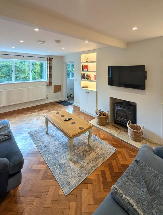 The living room with its wood burner, coffee table on a blue rug, and a window onto greenery