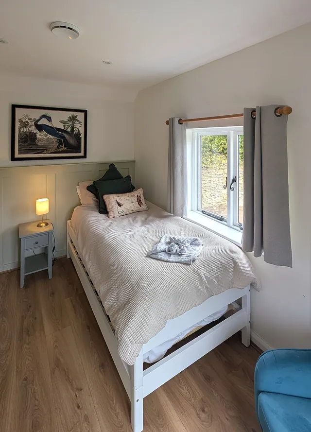 The second bedroom, set up with one single bed, with a heron print above, a bedside lamp and a window onto a Cotswold stone wall